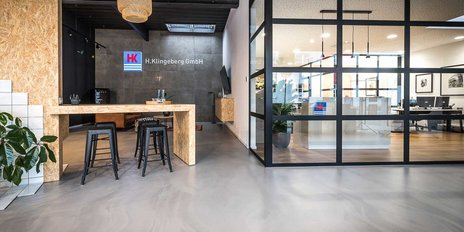 Ein moderner Empfangsbereich mit einem großen Holztisch und schwarzen Barhockern, umgeben von Pflanzen und Glaswänden.