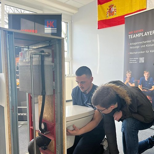 Zwei junge Männer arbeiten zusammen an einer Toilette in einem Schulungsraum, um praktische Fähigkeiten zu erlernen.
