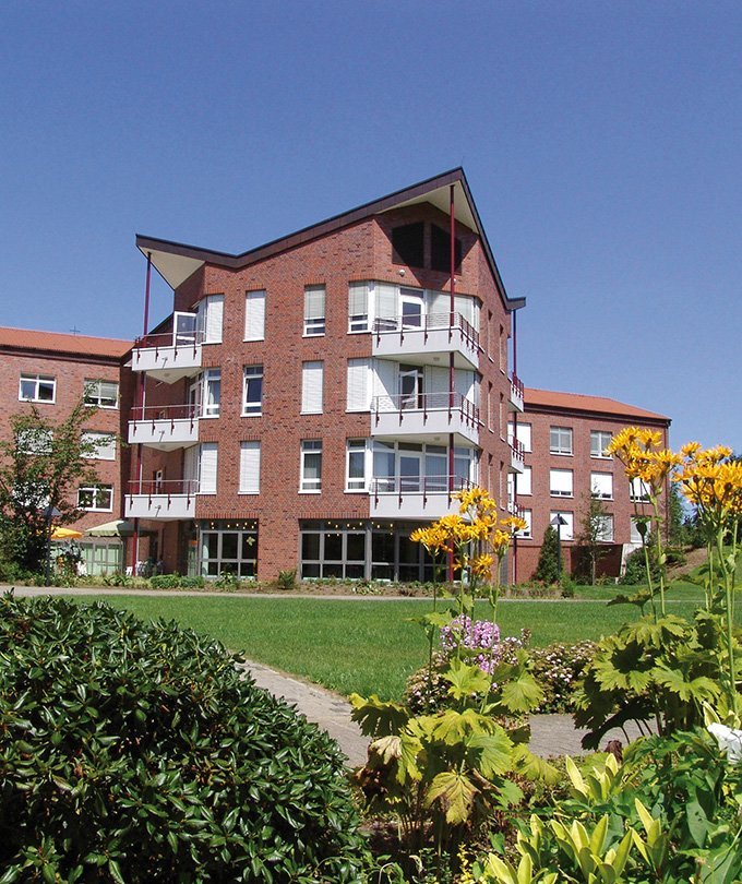 Ein modernes, mehrstöckiges Gebäude aus roten Ziegeln mit Balkonen und großen Fenstern, umgeben von blühenden Pflanzen.