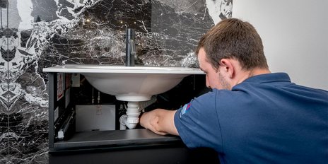 Ein Handwerker beugt sich über ein Waschbecken und arbeitet an den Rohren unterhalb, um ein Problem zu beheben.