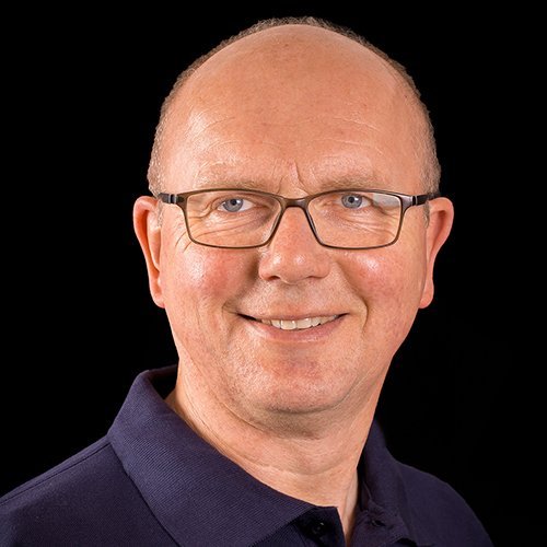 Ein lächelnder Mann mit Brille trägt ein dunkelblaues Poloshirt vor einem schwarzen Hintergrund.