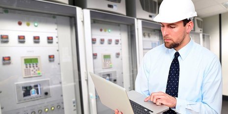 Ein Mann mit Bauhelm und einem Laptop steht in einem Serverraum