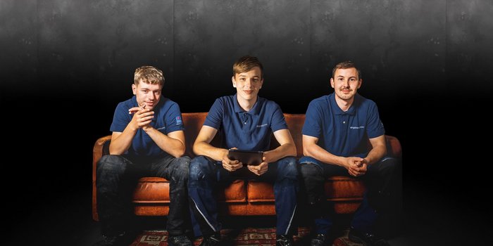 Drei junge Männer in blauen Poloshirts sitzen entspannt auf einer braunen Couch vor einem dunklen Hintergrund.