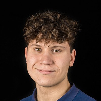 Ein junger Mann mit lockigem, braunem Haar lächelt freundlich in die Kamera vor einem schwarzen Hintergrund.
