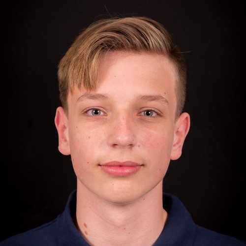 Ein junger Mann mit blonden Haaren und einem neutralen Gesichtsausdruck blickt direkt in die Kamera vor einem dunklen Hintergrund.