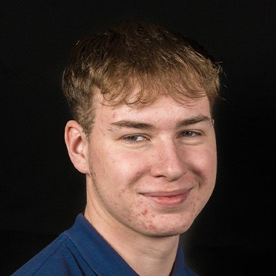 Ein junger Mann mit kurzen, blonden Haaren lächelt freundlich in die Kamera, während er ein blaues Hemd trägt.