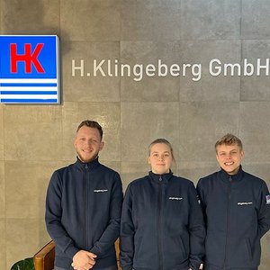 Drei Mitarbeiter stehen lächelnd vor dem Firmenlogo von H. Klingeberg GmbH in einem modernen Büro.