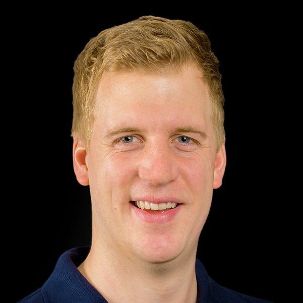 Der Mann hat kurze, blonde Haare und trägt ein dunkelblaues Hemd, während er direkt in die Kamera schaut.