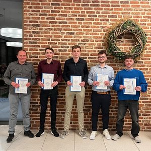 Fünf junge Männer stehen in einer Reihe vor einer Backsteinwand und halten Urkunden in den Händen.