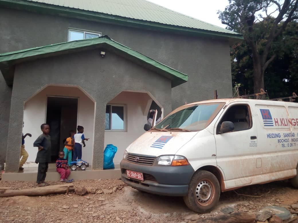 Ein graues Haus mit grünem Dach, vor dem Kinder spielen und ein Lieferwagen mit Firmenlogo parkt.