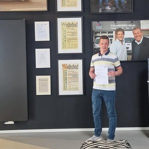 Der Mann in gestreiftem Shirt lächelt, während er stolz ein wichtiges Dokument vor einer Wand mit Auszeichnungen präsentiert.