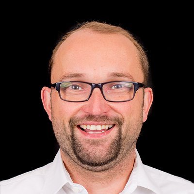 Der Mann zeigt ein breites Lächeln, seine Brille reflektiert das Licht, während er in die Kamera schaut.