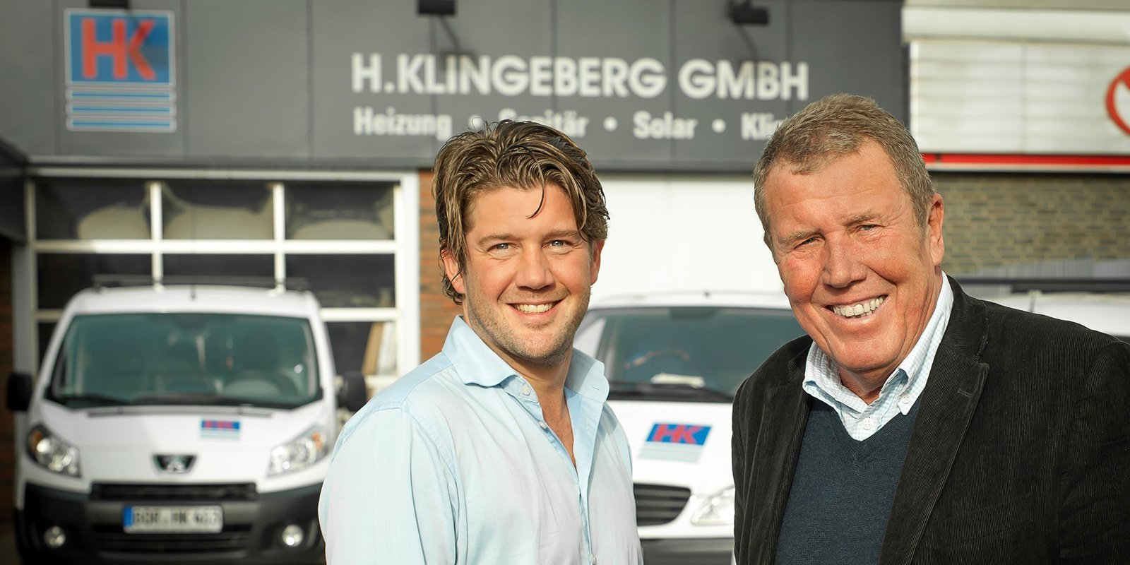 Zwei Männer stehen lächelnd vor dem Firmengebäude von H. Klingeberg GmbH, umgeben von Lieferwagen.