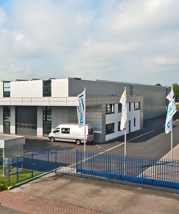 Ein modernes Firmengebäude mit großen Fenstern und einem grauen Lieferwagen, umgeben von blauen Fahnen und einem Zaun.