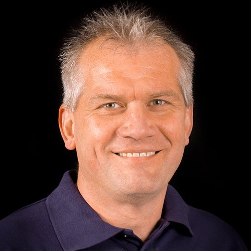 Ein lächelnder Mann mit grauen Haaren trägt ein dunkelblaues Polohemd vor einem schwarzen Hintergrund.