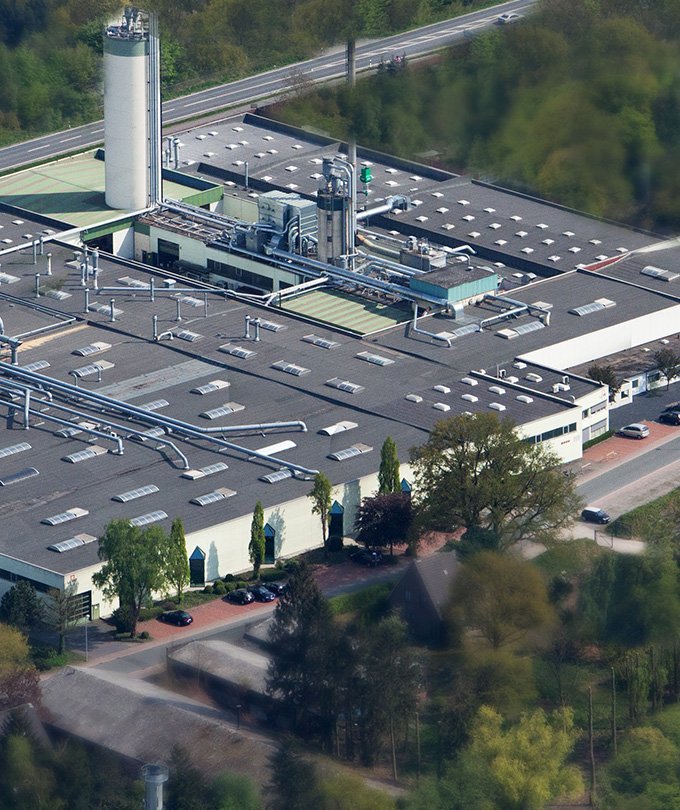 Eine Luftaufnahme einer großen Fabrik mit mehreren Rohren und einem hohen Schornstein, umgeben von Bäumen und einer Straße.