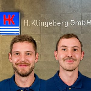 Zwei Männer in blauen Poloshirts lächeln vor einer Wand mit dem Logo der H. Klingeberg GmbH.