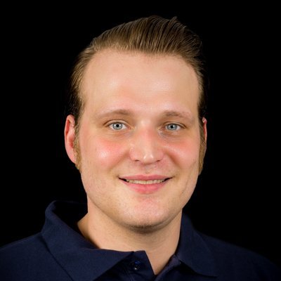 Ein junger Mann mit blonden Haaren und einem freundlichen Lächeln trägt ein dunkelblaues Hemd vor einem schwarzen Hintergrund.