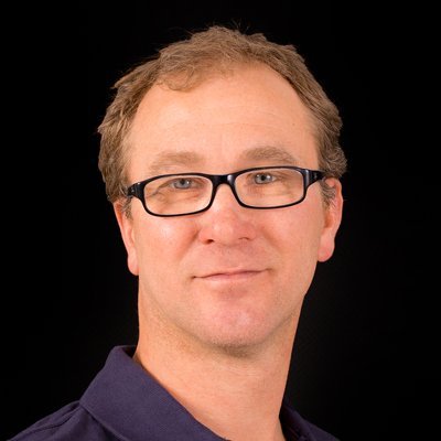 Ein Mann mit Brille und kurzem, lockigem Haar lächelt leicht vor einem schwarzen Hintergrund.
