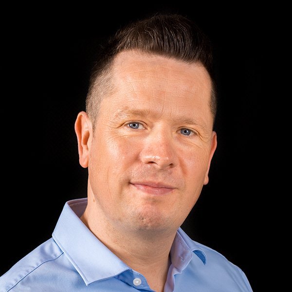 Ein Mann mit kurzem, gestyltem Haar und einem freundlichen Gesichtsausdruck trägt ein hellblaues Hemd vor einem schwarzen Hintergrund.