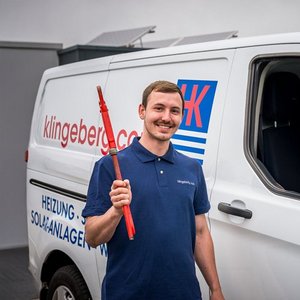 Ein junger Mann in einem blauen Poloshirt steht vor einem weißen Lieferwagen und hält ein rotes Werkzeug in der Hand.