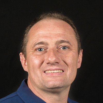 Ein Mann mit kurzen, braunen Haaren und einem freundlichen Lächeln trägt ein blaues Hemd vor einem dunklen Hintergrund.