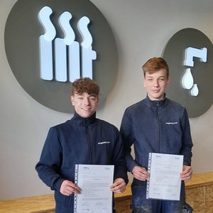 Zwei junge Männer stehen lächelnd nebeneinander und halten Zertifikate in der Hand, vor einer modernen Wandgestaltung.