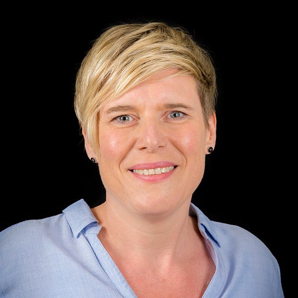 Eine lächelnde Frau mit kurzen blonden Haaren trägt ein hellblaues Hemd und hat einen freundlichen Ausdruck im Gesicht.