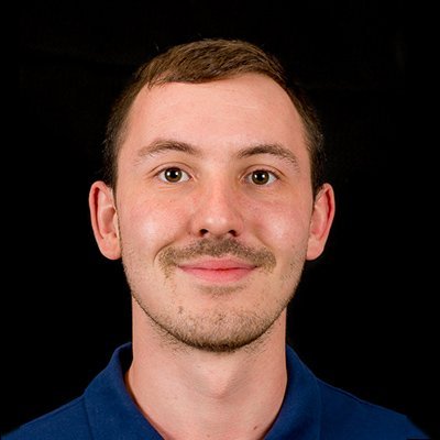 Der Mann hat einen neutralen Gesichtsausdruck und schaut direkt in die Kamera, während er ein blaues Polohemd trägt.