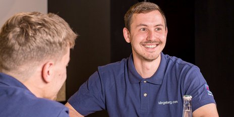 Ein junger Mann mit einem breiten Lächeln sitzt an einem Tisch und scheint sich angeregt zu unterhalten.