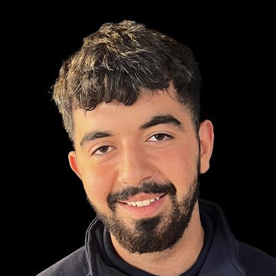 Der Mann trägt einen dunklen Pullover und hat einen gepflegten Bart, während er selbstbewusst in die Kamera lächelt.