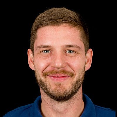 Ein junger Mann mit einem freundlichen Lächeln und einem kurzen Bart trägt ein blaues Hemd vor einem schwarzen Hintergrund.