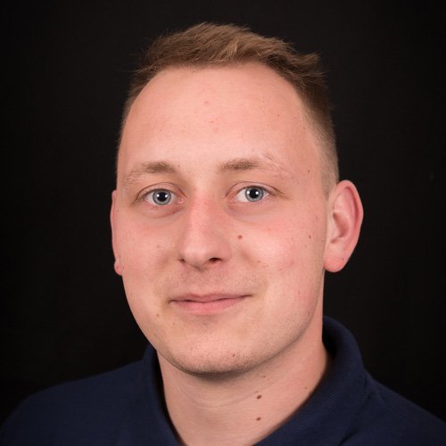 Ein junger Mann mit kurzen, blonden Haaren und blauen Augen lächelt freundlich vor einem dunklen Hintergrund.