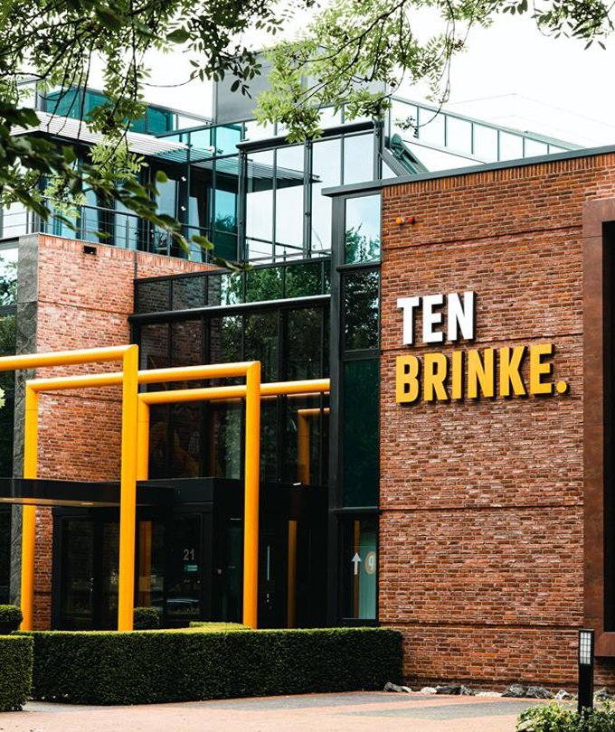 Das moderne Gebäude mit einer Backsteinfassade und großen Fenstern hat eine auffällige gelbe Beschilderung mit dem Namen 'TEN BRINKE'.
