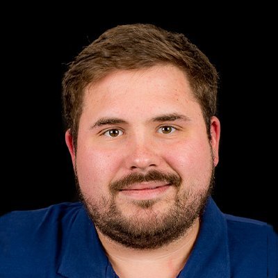 Ein junger Mann mit einem dichten Bart und einem freundlichen Lächeln trägt ein blaues Hemd vor einem schwarzen Hintergrund.