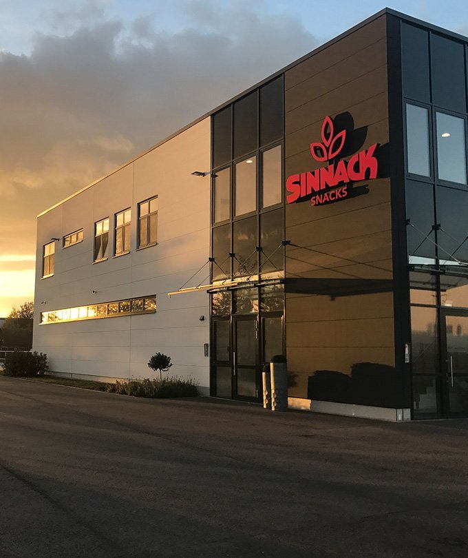Das moderne Gebäude mit großen Fenstern und dem roten Logo von Sinnack Snacks strahlt in der Abendsonne.