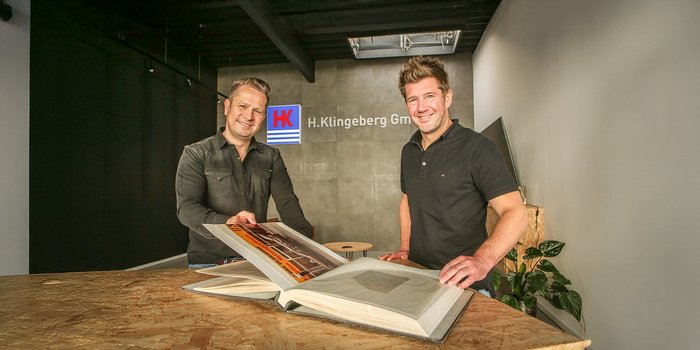 Zwei Männer stehen an einem Tisch und blättern in einem großen Buch, während sie lächeln und sich unterhalten.