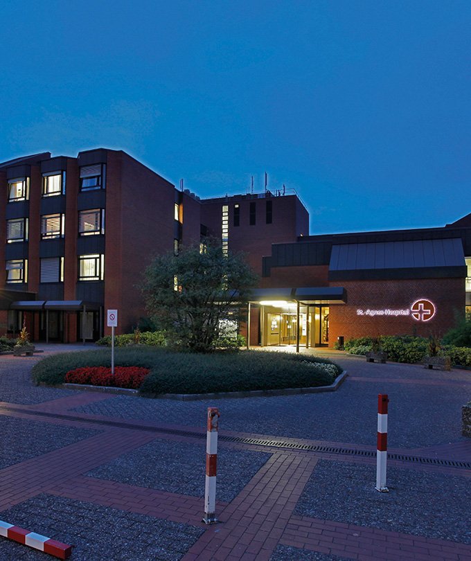 Das St. Agnus Krankenhaus erstrahlt in der Dämmerung, umgeben von gepflegten Gärten und einem einladenden Eingangsbereich.