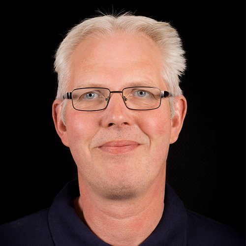 Ein lächelnder Mann mit Brille und grauen Haaren, der in einem dunklen Hemd vor einem schwarzen Hintergrund steht.