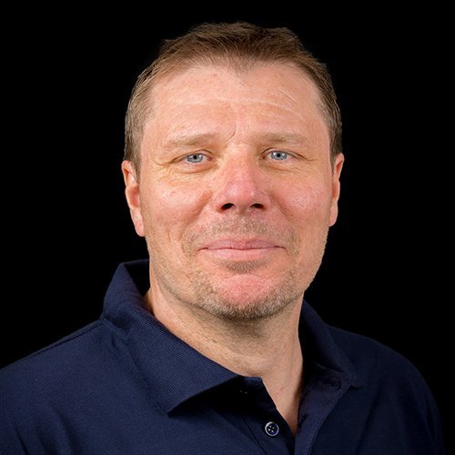 Ein Mann mit kurzen, blonden Haaren und blauen Augen lächelt freundlich in die Kamera, während er ein dunkelblaues Polohemd trägt.