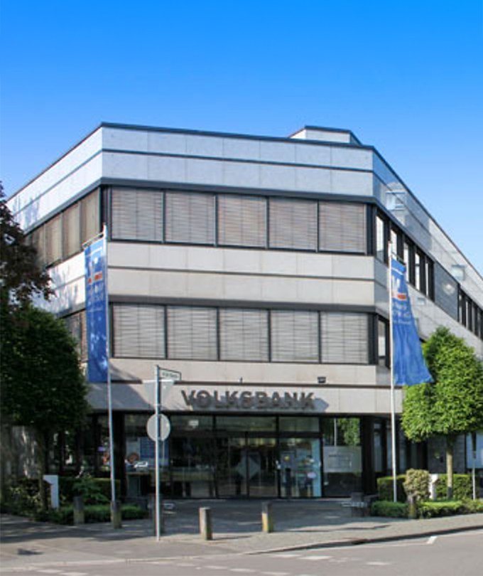 Das moderne Gebäude der Volksbank mit großen Fenstern und einem klaren blauen Himmel im Hintergrund ist gut sichtbar.