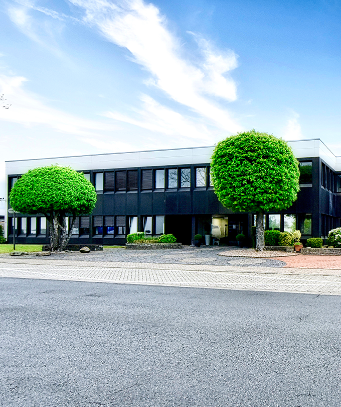 Ein modernes Bürogebäude mit großen Fenstern, umgeben von gepflegten Grünflächen und runden, buschigen Bäumen.