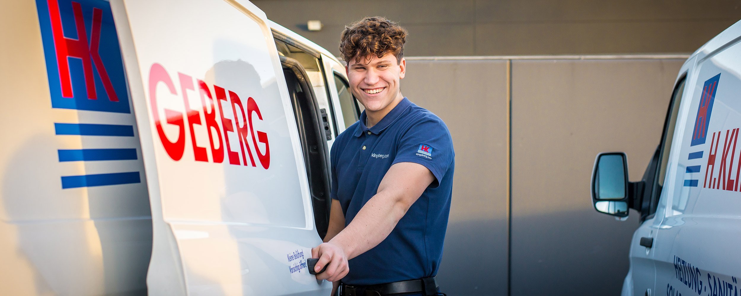Ein junger Mann mit lockigem Haar lächelt, während er an einem Lieferwagen steht, der mit 'H. Geberg' beschriftet ist.