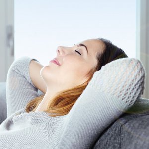 Eine Frau entspannt sich auf einem grauen Sofa, mit geschlossenen Augen und einem zufriedenen Lächeln im Gesicht.