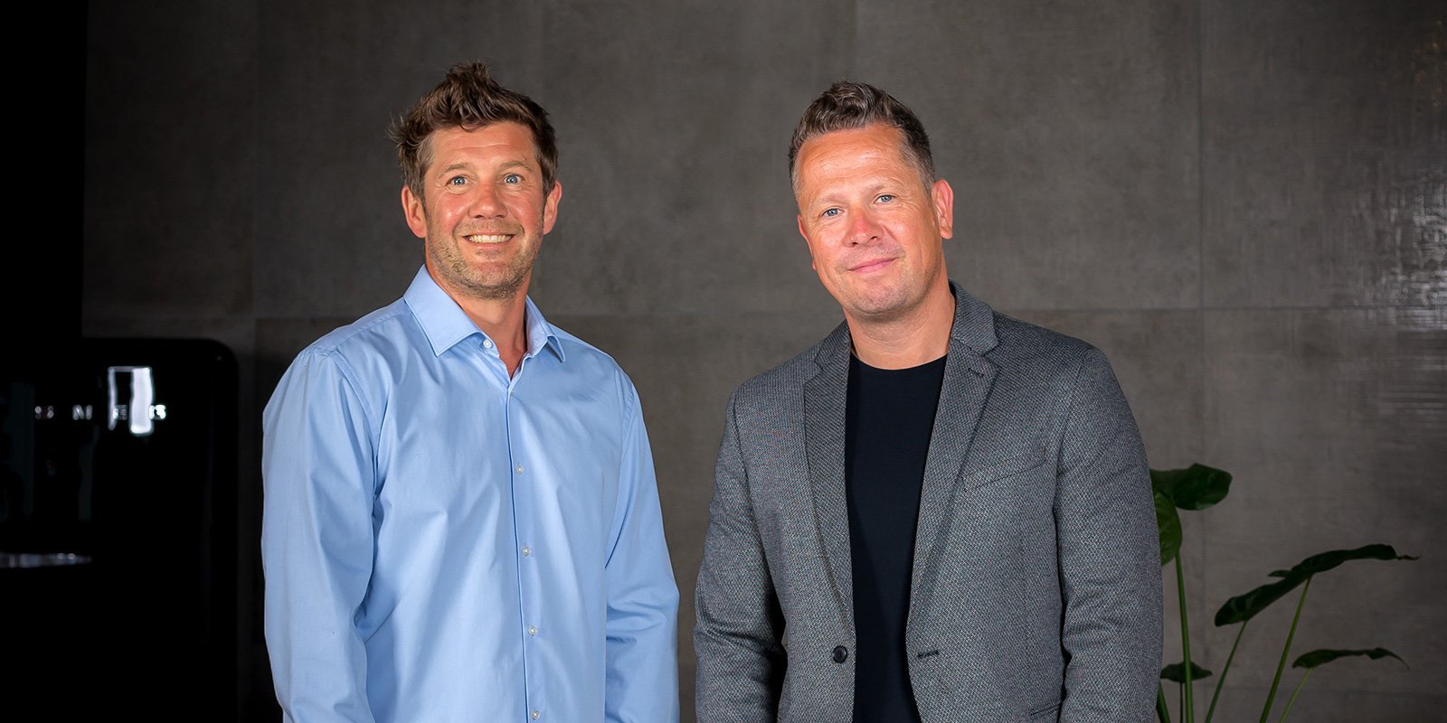 Zwei Männer stehen lächelnd nebeneinander in einem modernen Raum mit grauen Wänden und einer Pflanze im Hintergrund.