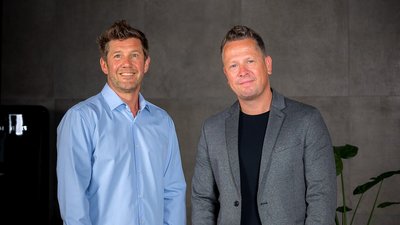 Zwei Männer stehen lächelnd nebeneinander in einem modernen Raum mit grauen Wänden und einer Pflanze im Hintergrund.