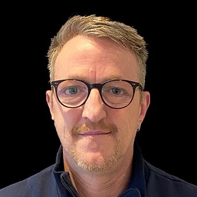 Ein Mann mit Brille und kurzem, grauem Haar blickt direkt in die Kamera, mit einem neutralen Gesichtsausdruck.