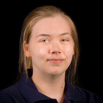 Eine junge Frau mit blonden Haaren und einem neutralen Gesichtsausdruck trägt ein dunkles Oberteil vor einem schwarzen Hintergrund.