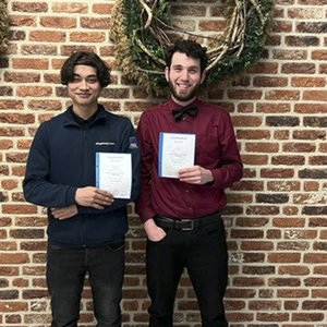Zwei junge Männer stehen vor einer Backsteinwand und halten stolz ihre Zertifikate in den Händen.