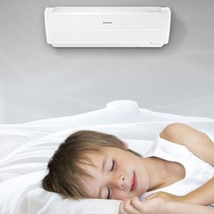 Ein schlafendes Kind liegt in einem hellen Bett, während eine Klimaanlage über ihm an der Wand montiert ist.